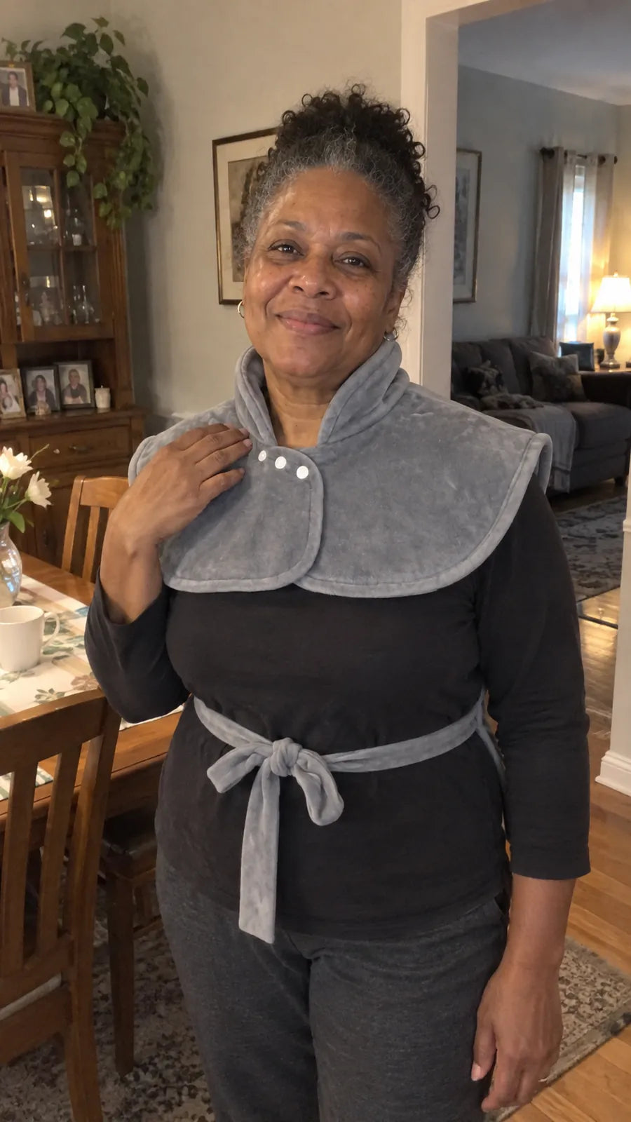 A smiling woman in her dining room wearing the Heatcape wrap, collar snapped and waist tie knotted.